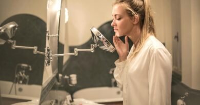 Side view of blond woman in white blouse standing in bathroom looking closely at face skin in small mirror