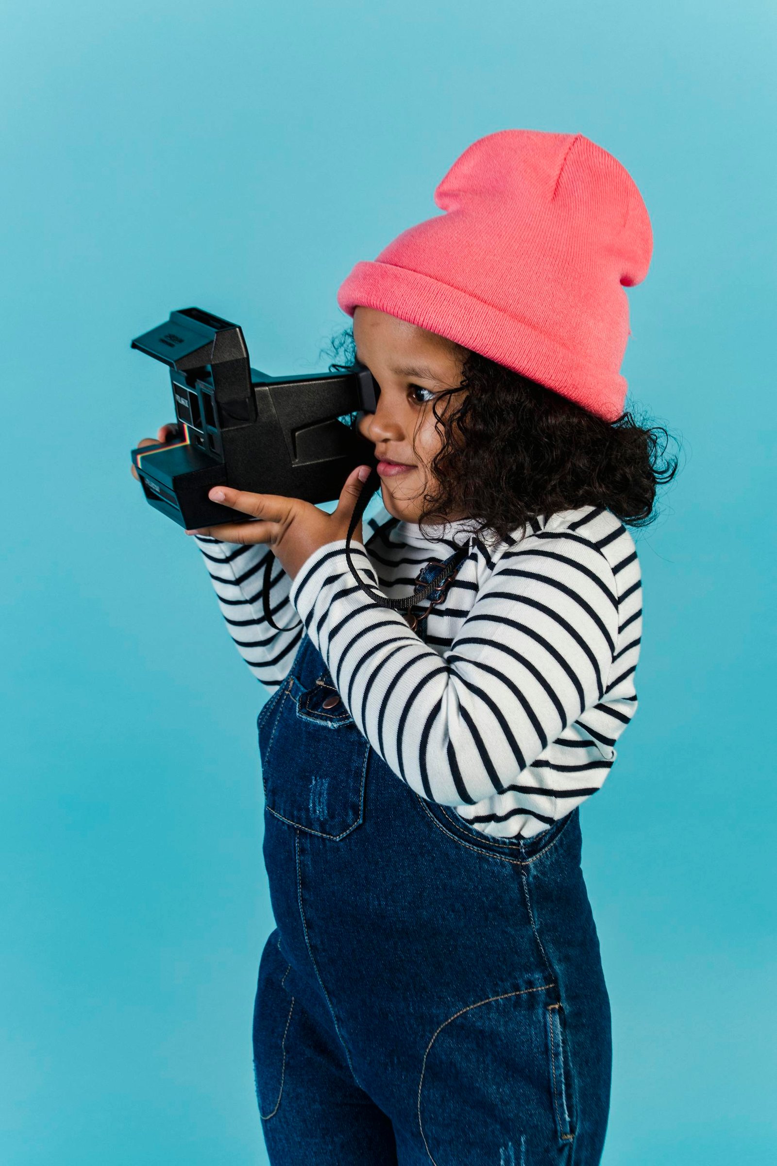 Curious black girl with curly hair in stylish clothes taking photo on instant retro photo camera against blue background in studio
